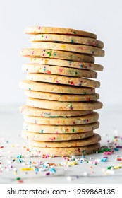 Close Up Of A Stack Of Homemade Sprinkle Sugar Cookies Against A Light Background With Sprinkles Scattered Around.