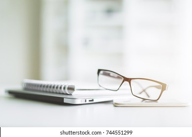 Close up of stacked laptop computer, glasses, spiral notepad and smartphone on blurry office table - Powered by Shutterstock