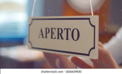 Close Up Of Staff Turning Aperto Sign Hanging On Glass Door In Italian Restaurant. Owner Or Seller Turning Open Sign Written In Itaian On Cafe Window