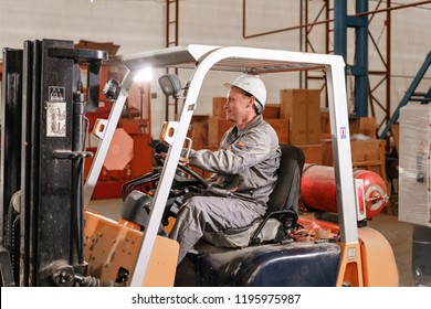 Close - Up Steering Wheel And Levers. Man Driving A Forklift Through A Warehouse In A Factory. Driver In Uniform And Protective Helmet. The Concept Of Logistics And Storage