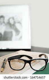 Close Up Still Life Of A Pair Of Reading Glasses And A Black Pen Laying On An Orange Financial Newspaper On A Dark Wooden Writing Desk With A Family Photo Frame. Office Interior With No People.