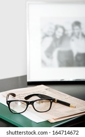 Close Up Still Life Of A Pair Of Reading Glasses And A Black Pen Laying On An Orange Financial Newspaper On A Dark Wooden Writing Desk With A Family Photo Frame. Office Interior With No People.