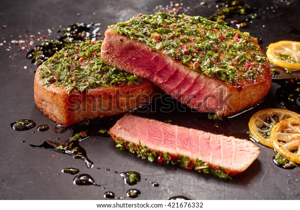 Close Up\
Still Life of Seared Tuna Steaks Topped with Fresh Pesto on Dark\
Gray Surface Dirtied with Oil and Lemon\
Slices