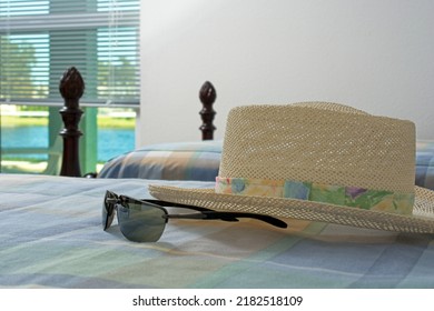 Close Up Of Straw Hat And Sunglasses Lying On Bed In Room With Window Overlooking Water Front View. Winter Break Holiday Scene In Florida. Copy Space.