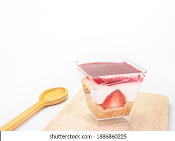 The close up of strawberry cheesecake in transparent plastic square shape cup with wood spoon on wooden plank on white background.