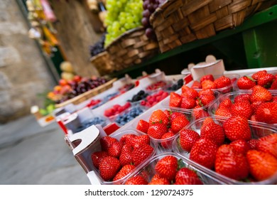 Close up of strawberry inside clear plastic box in open white paper box in fruit stall with many kind of fruits in wooden basket background, selective focus
