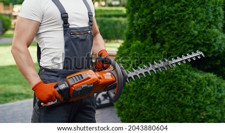 Close up of strong male gardener in brown overalls and gloves holding electric hedge trimmer while standing on back yard. Concept of people and landscaping.