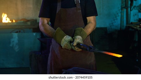 Close Up Of Strong Man In Safety Apron And Gloves Holding Pliers With Hot Steel. Competent Blacksmith At Workplace. Hard Manual Work At Forge.