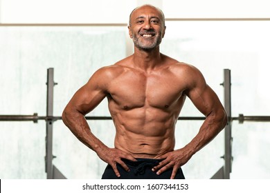 Close up of  strong muscular shirtless mature older bodybuilding athlete with balding gray hair  with hands on hips smiling at camera in a gym