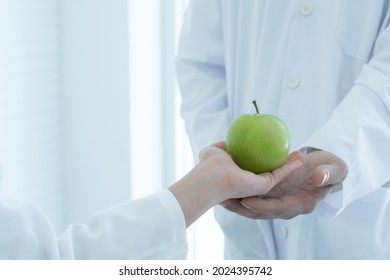 Close Up Of Student Hand Wears Lab Coat Giving Green Apple To Science Teacher In Lab, Selective Focus