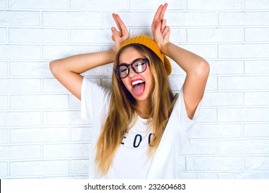 Close up studio portrait of cheerful blonde hipster girl going crazy making funny face and showing her tongue. White urban wall background. 