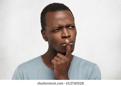 Close Up Studio Portrait Of Handsome Young Black Man Having Concentrated Thoughtful Expression, Frowning, Keeping Finger On His Lips As If Trying To Remember Something Or Saying: And What If.