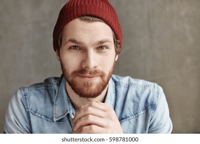 Close up studio portrait of stylish brutal young bearded European man wearing hat and jeans jacket having rest indoors, posing at blank wall with copy space for your text or promotional content