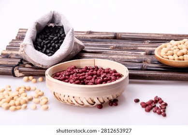 A close up studio shot of assorted raw beans and legumes, including red adzuki beans in a bamboo bowl, black beans in a small sack, and soybeans scatt
