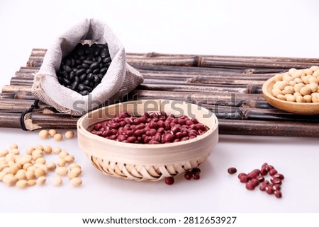A close up studio shot of assorted raw beans and legumes, including red adzuki beans in a bamboo bowl, black beans in a small sack, and soybeans scatt