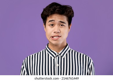 Close up studio shot of young Asian male making silly face, grimacing from the unpleasant sensation or sound looking at camera, posing at purple violet wall. Human facial expressions and emotions