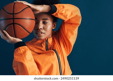 Close up studio shot of young female basketball player in orange sportswear posing with ball. Portrait of a professional basketball player over blue background. - Powered by Shutterstock