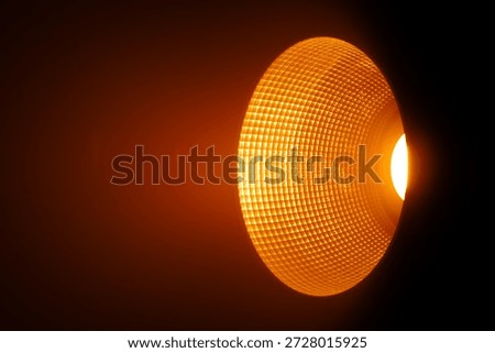 Close up of a studio spotlight emitting warm orange light with a textured reflector in a dark surrounding space. Lighting equipment backstage.