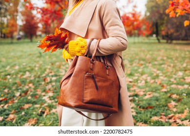 Close up of stylish leather handbag held by woman wearing coat in autumn park. Fall female clothes, accessories. Fashion