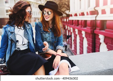 Close up  summer lifestyle portrait of two best friends laughing and talking  outdoor on the street in city center . Wearing stylish jeans jacket , dress, sunglasses. Enjoying time together.