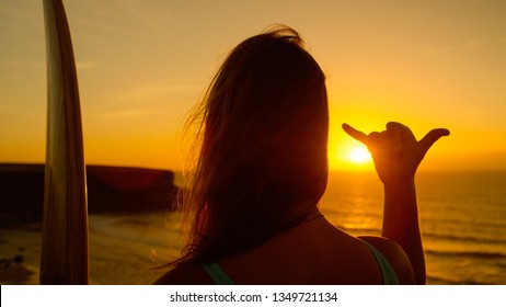 CLOSE UP SUN FLARE: Cool Surfer Girl Gives The Shaka Sign While Watching The Summer Sunset. Unrecognizable Woman Holding Her Surfboard Shows A Hang Loose Gesture With Her Hand Over The Golden Sun.