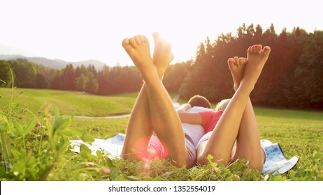 CLOSE UP SUN FLARE: Unrecognizable barefoot man and woman lie in the serene countryside and enjoy a romantic date. Young boyfriend and girlfriend lying embraced on a blanket in the sunlit meadow. - Powered by Shutterstock