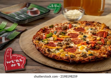 Close Up Of Supreme Pizza With Cauliflower Crust Topped With Sausage, Pepperoni, Peppers, Onions, Zucchini, Cheese And Tomato Sauce Sitting On Pizza Peel Surrounded By Football Themed Decorations