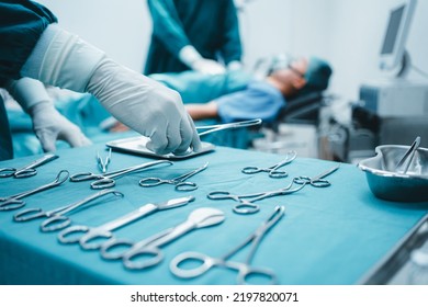 Close Up Surgeon Doctor's Hand With Hygiene Glove Taking Sterile Surgical Instrument Tool, Equipment In Operating Room Patient In Background For Surgery. Hospital, Medical Healthcare Emergency Concept