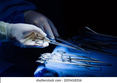 close up surgeons hand taking scissors, forceps and surgical instruments on table for operation with colleagues performing work in operation room at hospital, emergency case, surgery, medical concept
