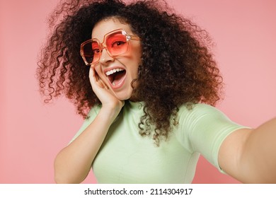 Close Up Surprised Young Curly Latin Woman 20s Wears Mint T-shirt Sunglasses Doing Selfie Shot Pov On Mobile Phone Put Palm Hand On Face Isolated On Plain Pastel Light Pink Background Studio Portrait