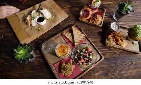 Close up of table in restaurant served with assorted food on wooden platters. Image of different dishes and snacks on the brown surface with people and glasses of red wine in the background. - Powered by Shutterstock