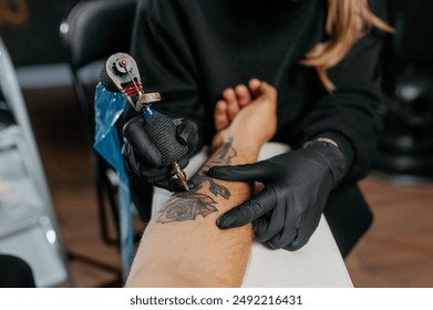 Close up tattoo master  professional tattoo artist in black gloves making a  rose tattoo on a man's forearm. He is holding  a tattoo machine. - Powered by Shutterstock