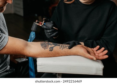 Close up tattoo master  professional tattoo artist in black gloves making a  rose tattoo on a man's forearm. He is holding  a tattoo machine.