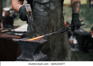 Close up of tattooed incognito blacksmith manually forging molten metal on anvil outdoors. Male blacksmith in leather apron and black gloves working with hammer. Concept of blacksmithing.