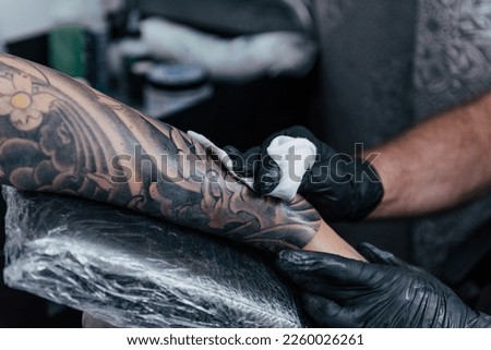Close up tattooist cleaning fresh tattoo. Final process on forearm with white ink. Detail black gloves tattooing. Tattoo artist working in studio. Creative small business and tattoo master.