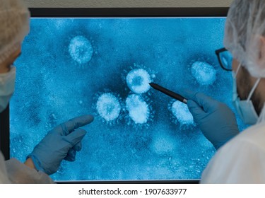 Close up of team of scientists in masks and medical gloves sits at computer monitor in laboratory and discusses photo of Coronovirus under microscope. COVID-19. 