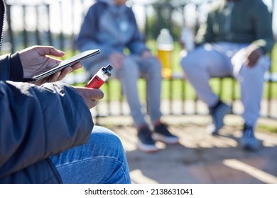 Close Up Of Teenagers With Mobile Phone Vaping And Drinking Alcohol In Park