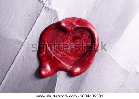 Close Up Texture of a Melted Red Sealing Wax Stamp in a Heart Shape Pressed onto a Shiny Metallic Silver Foil Background