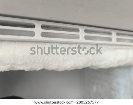 Close up of a thick layer of white ice and frost buildup on a domestic freezer shelf vent grill, heavy hoarfrost accumulation inside a refrigerator cooling unit needing manual defrosting