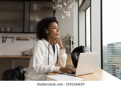 Close up thoughtful African American businesswoman looking out window, touching chin, sitting at wooden table with laptop and motorcycle helmet, pondering project strategy, planning, visualizing - Powered by Shutterstock