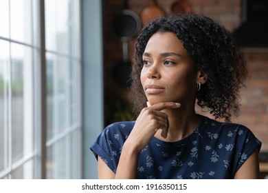 Close up thoughtful African American woman looking out window to aside, touching chin, dreamy young female lost in thoughts, planning, visualizing future, businesswoman thinking, making decision