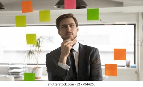 Close Up Thoughtful Businessman Touching Chin, Reading Notes On Colorful Stickers, Pensive Executive Boss Wearing Suit Pondering Corporate Project Strategy Or Work Plan, Standing Near Glass Wall