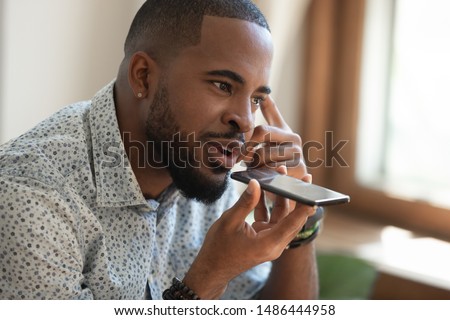 Close up thoughtful mindful african american guy sitting on couch, talking on speakerphone, dictating voice message, using online translator app or voice recognition software, virtual assistant.