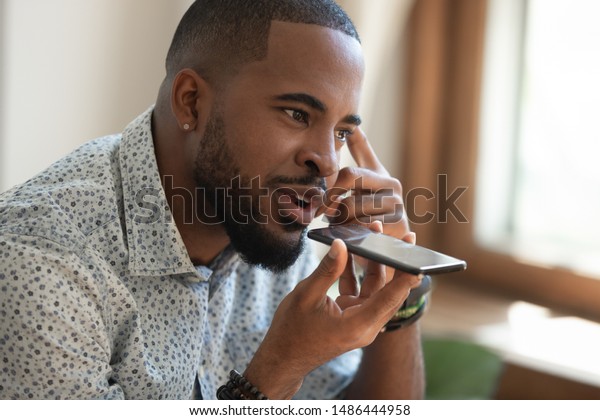 Close up thoughtful mindful african american\
guy sitting on couch, talking on speakerphone, dictating voice\
message, using online translator app or voice recognition software,\
virtual assistant.