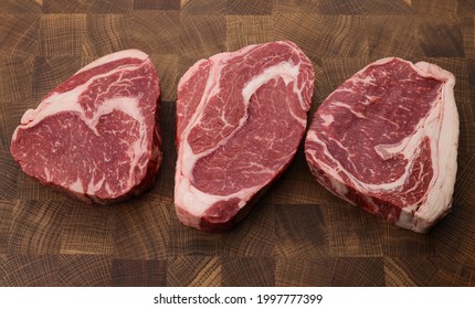 Close Up Three Raw Marbled Ribeye Beef Steaks Over End Grain Cutting Board Of Wooden Butcher Block, High Angle View, Directly Above