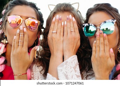 Close up of three young girls wearing in creative sunglasses with designed manicure, two lady covers mouth and one hiding face by hands. Happy friends posing and having fun together.
