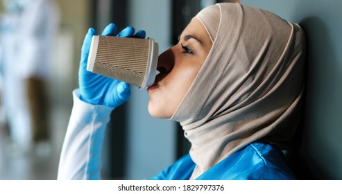 Close Up Of Tired Arab Young Woman Doctor In Hijab And Gloves Drinking Coffee And Leaning On Wall. Muslim Female Nurse In Headscarf Resting, Having Break, Sipping Drink. Coronavirus Work. Having Rest.