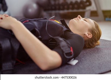 Close up of tired young woman in ems vest laying down with eyes close on mat in gym with weight rack in background