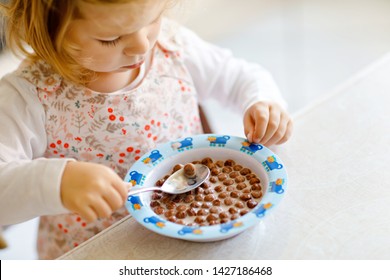 Close Up Of Toddler Girl Eating Healthy Cereal With Milk For Breakfast. Cute Happy Baby Child In Colorful Clothes Sitting In Kitchen And Having Fun With Preparing Oats, Cereals. Indoors At Home