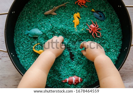 Close up of toddler hands playing with the blue rice in an ocean theme sensory tray play set up for toddler to explore
